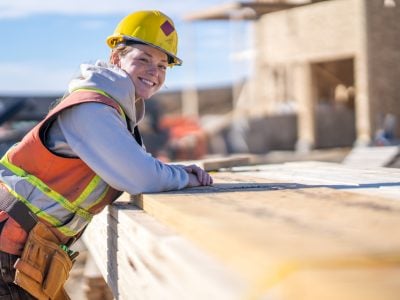 A female construction worker leans on a stack of lumber as she turns to pose for a portrait.  She is wearing proper safety equipment and is smiling warmly.